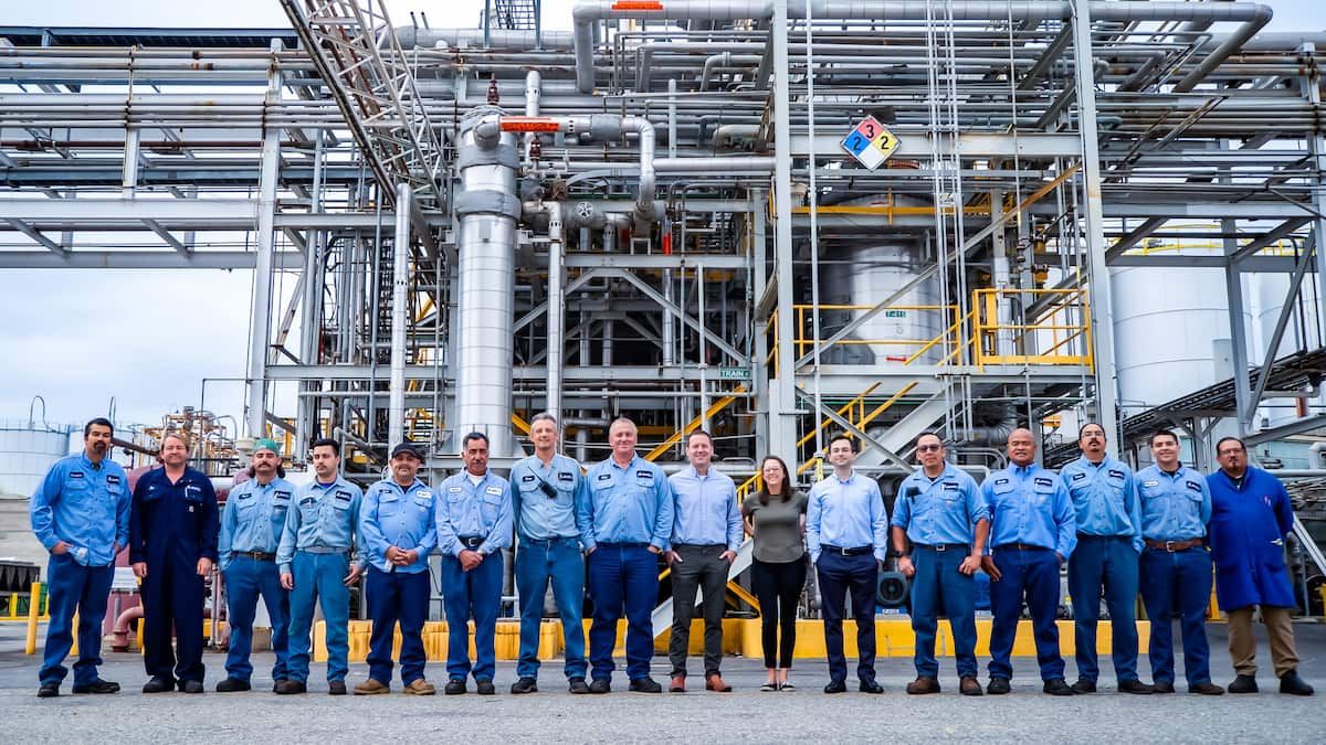 3 employees smile while wearing hard hats and hearing protection