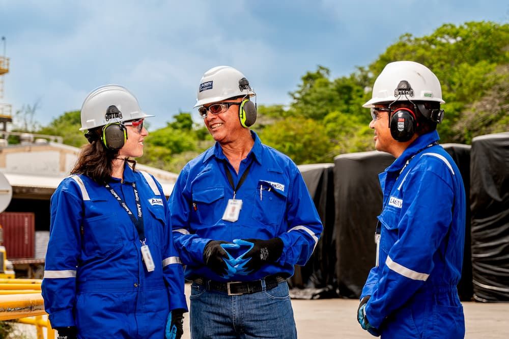 3 employees smile while wearing hard hats and hearing protection